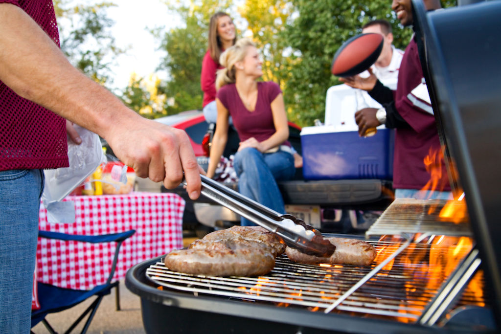 Man flipping hamburgers and hot dogs on grill with friends in background